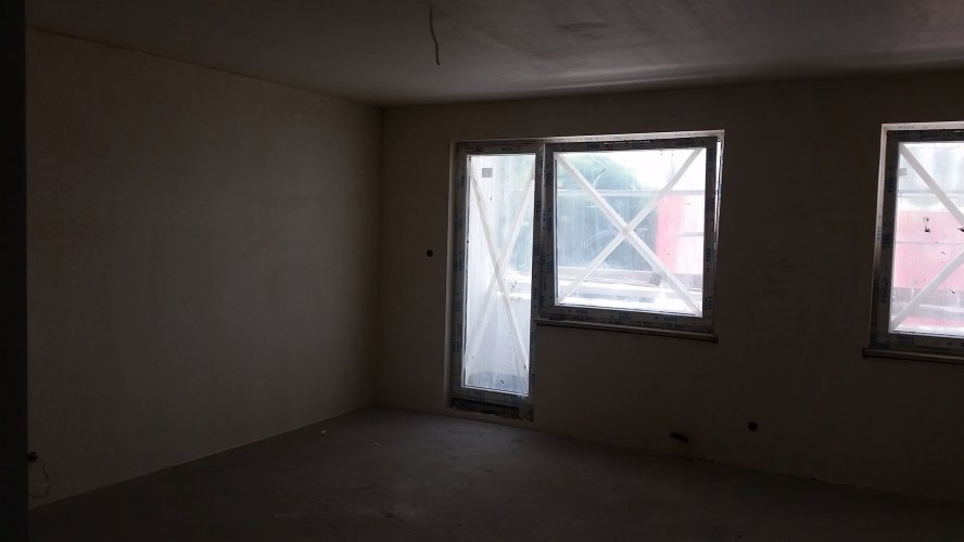Budynek Delta - wnętrze mieszkania po otynkowaniu, widoczne kwadratowe okna oraz drzwi prowadzące na duży balkon.