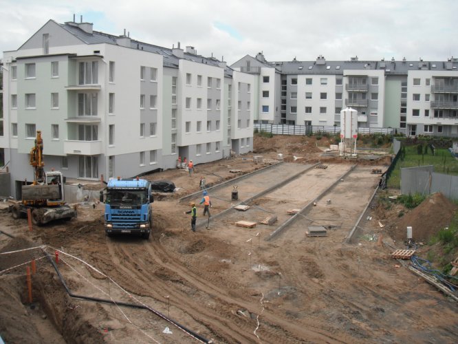 Teren osiedlowy przed budynkiem 16, widoczne prace przy realizacji drogi, chodników oraz miejsc parkingowych.