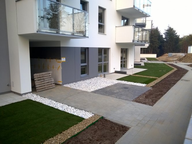 Budynek 1 - strefa wejścia do sekcji B, widoczne tarasy oraz trawniki w ogródkach mieszkań parterowych.